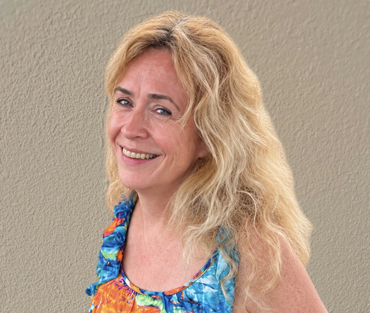 Smiling woman with long blonde hair in a colorful floral top against a beige wall background.